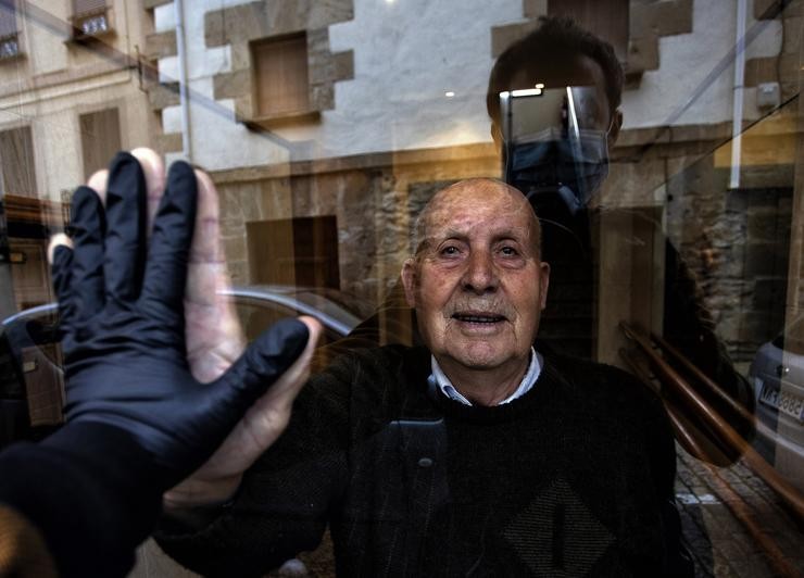 "Visita ao meu avó Miguel, de 90 anos, durante o confinamento, que saúda a través do cristal do portal,  Huarte, Navarra, 1 de abril, 2020". (© Unai Beroiz / Europa Press.