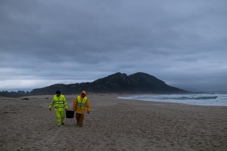 Dous homes cargan un cubo cheo de pellets, na praia Do Dique, a 13 de xaneiro de 2024, en Queiruga / Elena Fernández - Arquivo